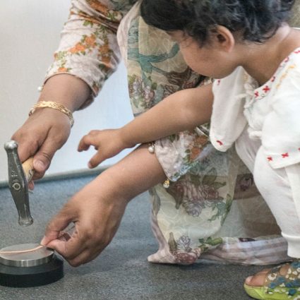 metal1 A very young girl reaches for a jewellers hammer being used by an older asian lady.