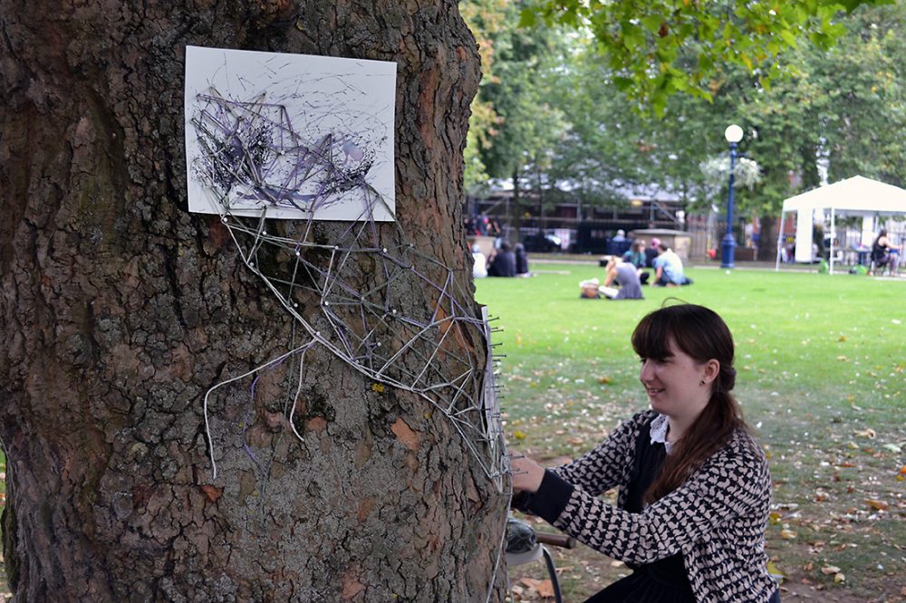 The artist places string around the pins placed on the tree. Ink drawings are placed underneath these.