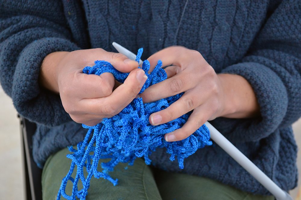 A pair of hands use a knitting needle to knit blue cord.