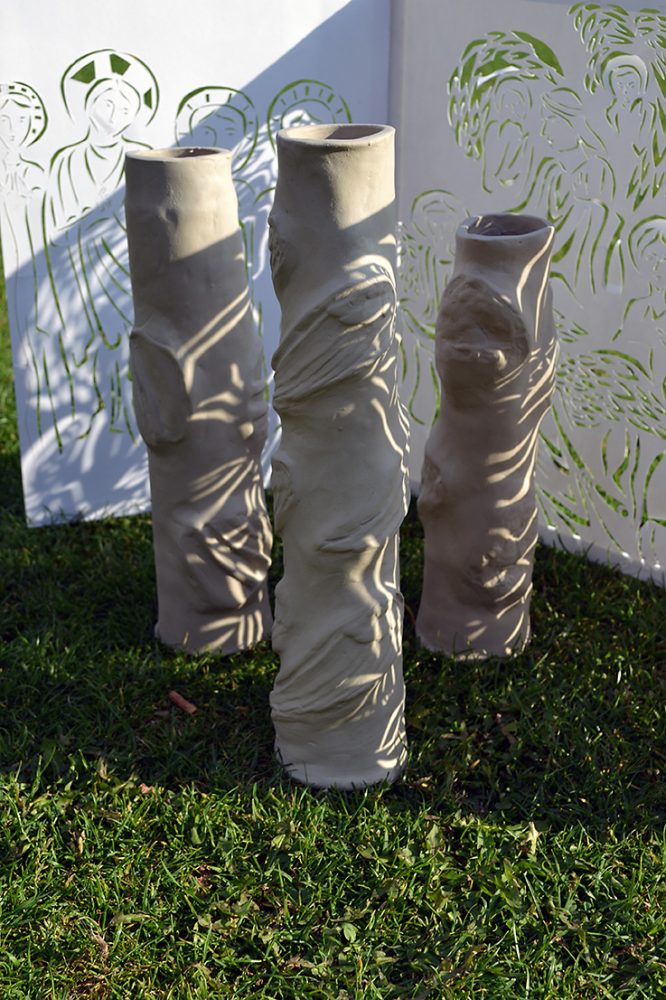 Three clay cylinders carved and manipulated into different shapes sit on the grass with a back drop of cut out paper showing figures from the bible.