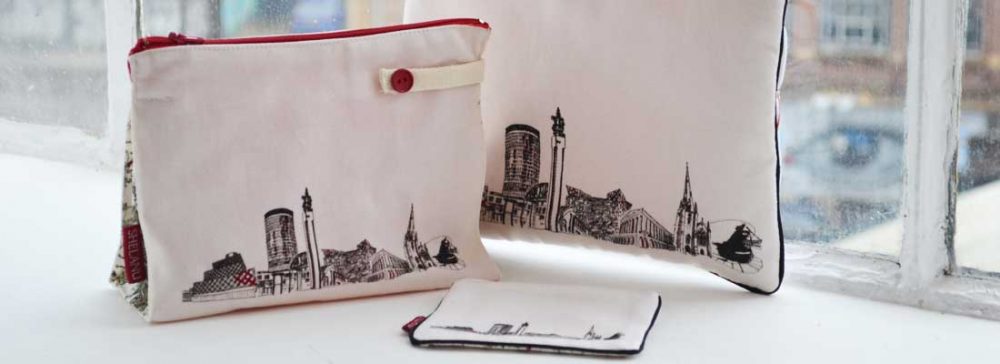 The i pad case, wash bag and purse sit on a windowsill. They are simple in design with a black and white line drawing of the skyline and red trimmings.