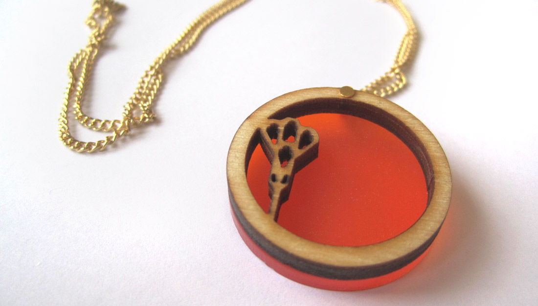 a gold necklace with a pendant made from a cut out circle of wood with a small section representing a bud. There is a disk of orange acrylic which the wood sits on top of.