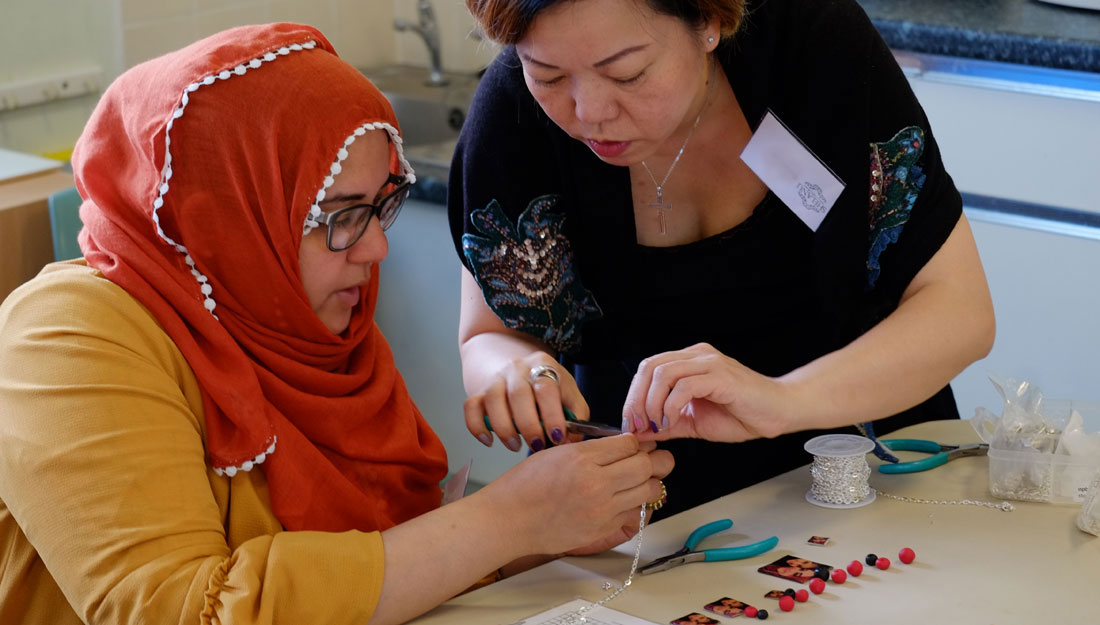 A woman helps another to attach a silver chain to a piece of jewellery. One woman wears a headscarf and the other woman is chinese.