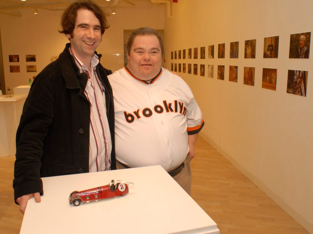 Two men smiling stand behind a plinth with a small model of a metal car on top of it.