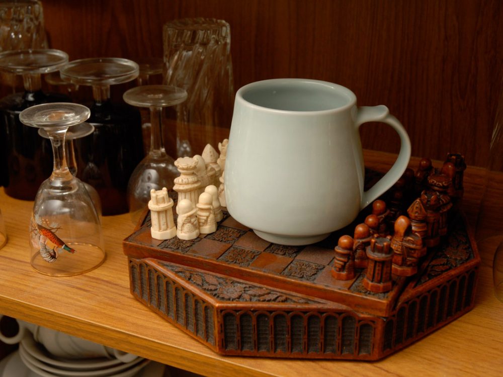 A white simple mug sits on an intricately carved chess board.