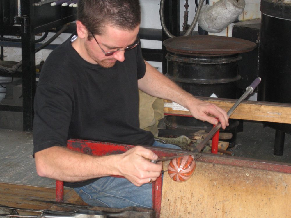 A glass blower uses his tools to cut the glass from a metal rod.