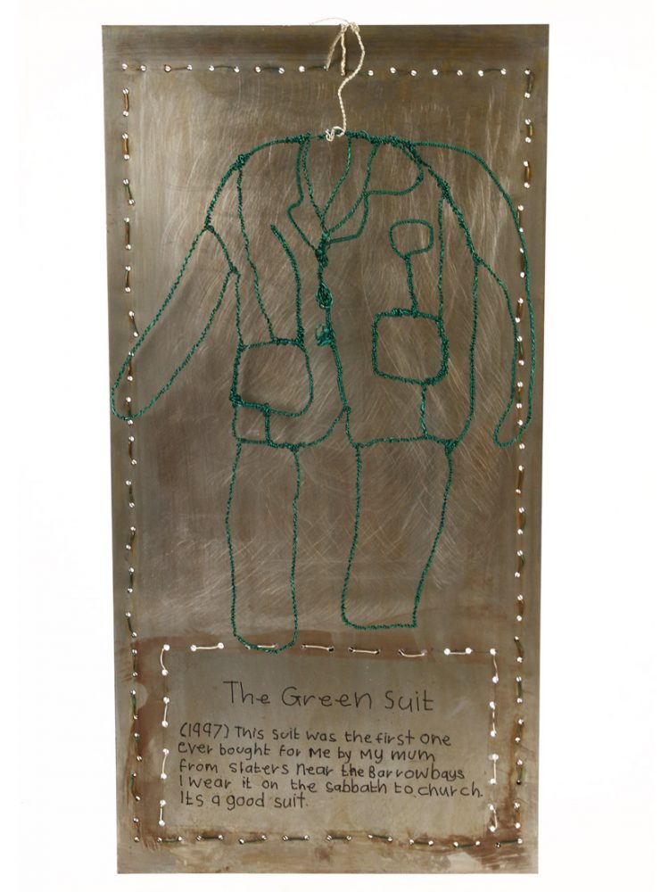 A metal plaque with text about a green suit. Green wire hangs infront of this in the shape of a suit.