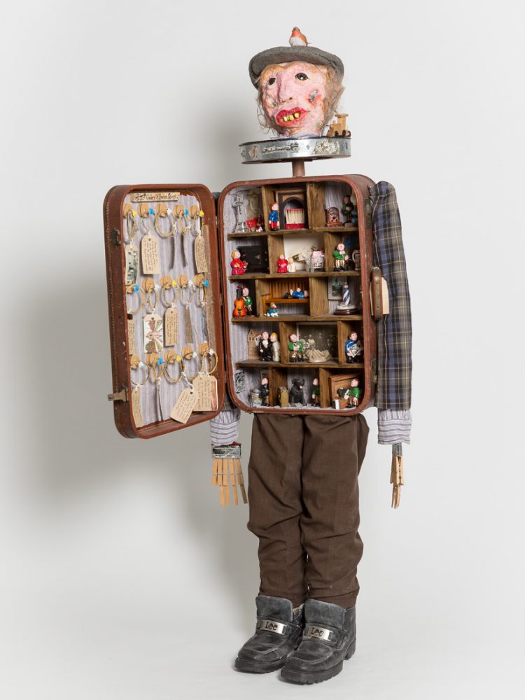 A figure made from found objects including pegs and a briefcase. The briefcase is open showing various figurines in differing settings.