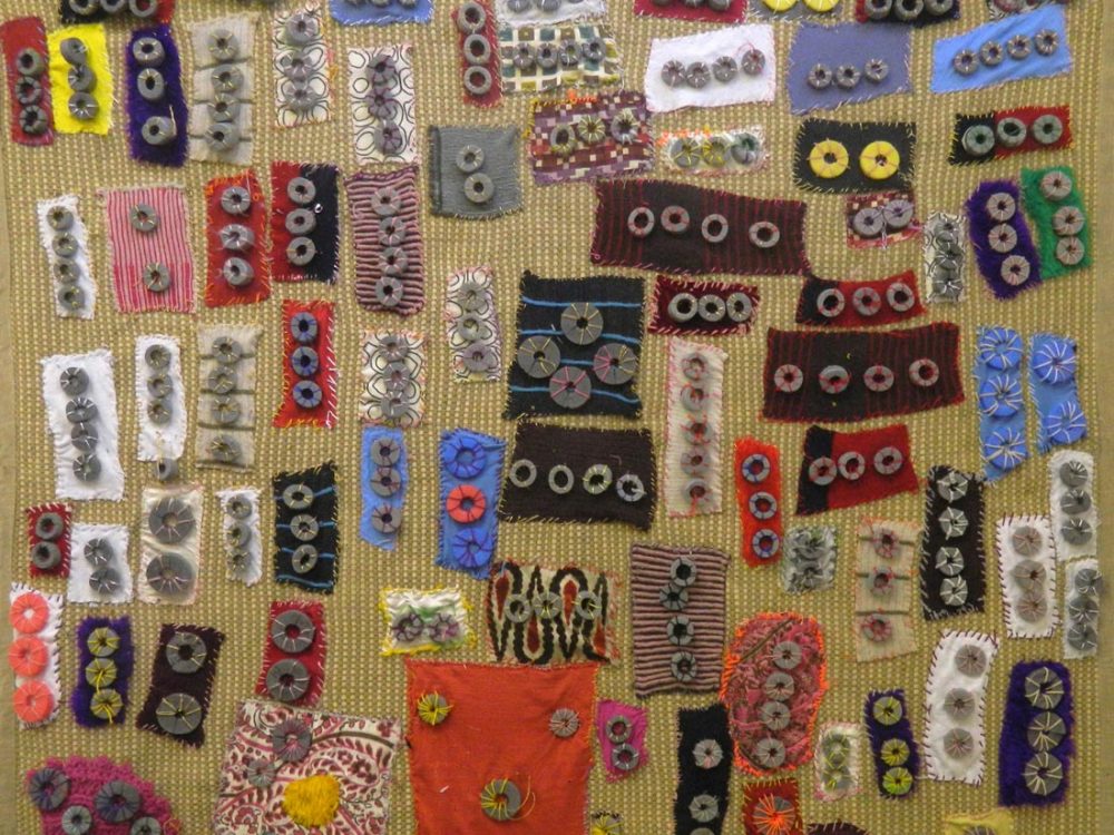 Close up of a rug with various patterned cloths stitched onto the surface with metal washers stitched on.