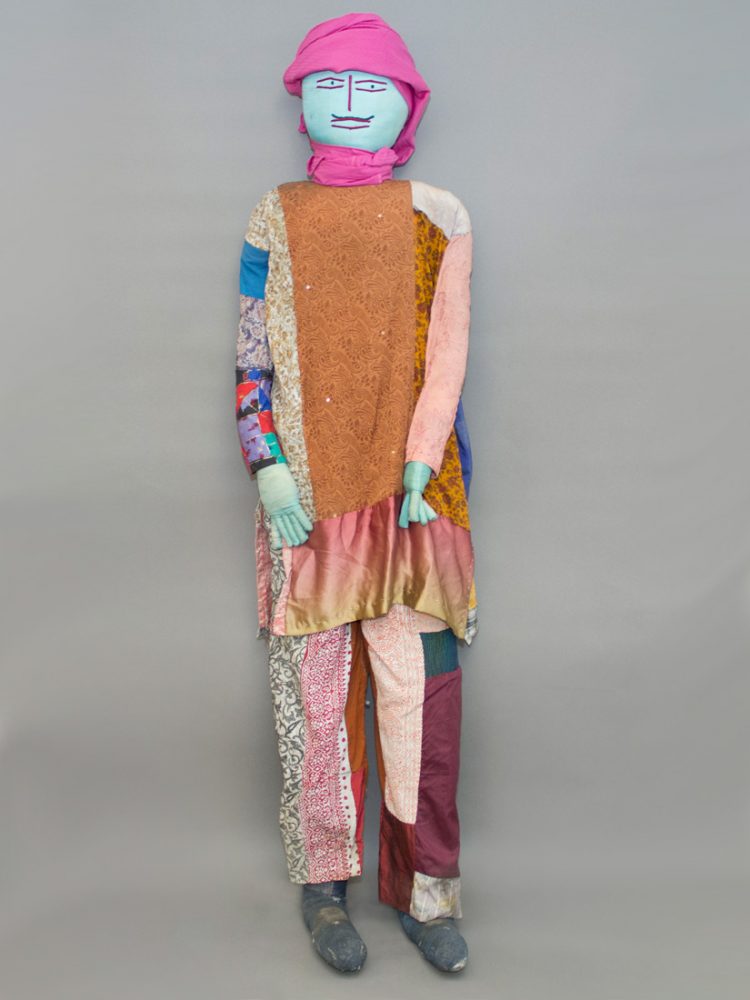 A 3D figure made from various pieces of cloth stands before a grey background.