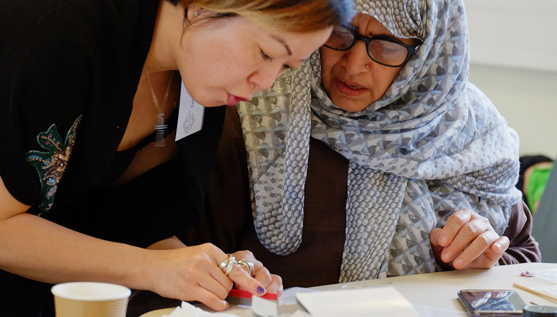 A woman demonstrates jewellery techniques to another older woman wearing glass and a headscarf.