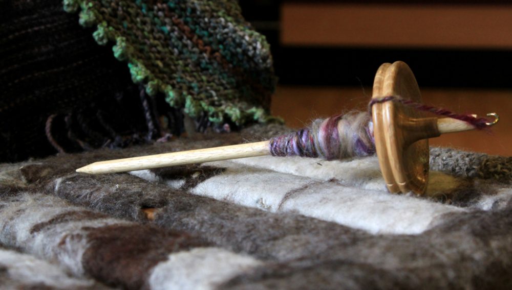 A drop spindle lies on a bench which has been covered with knitted pieces.