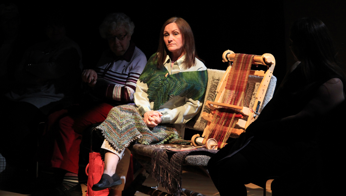 A woman sits on a bench, lit by a spotlight. A small loom next to her. She looks deep in thought.