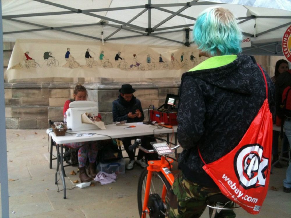 Outside, under a gazebo, a young man rides a stationary bike which is powering a sewing machine. A young woman sews and protraits of cyclists are hung around the space.