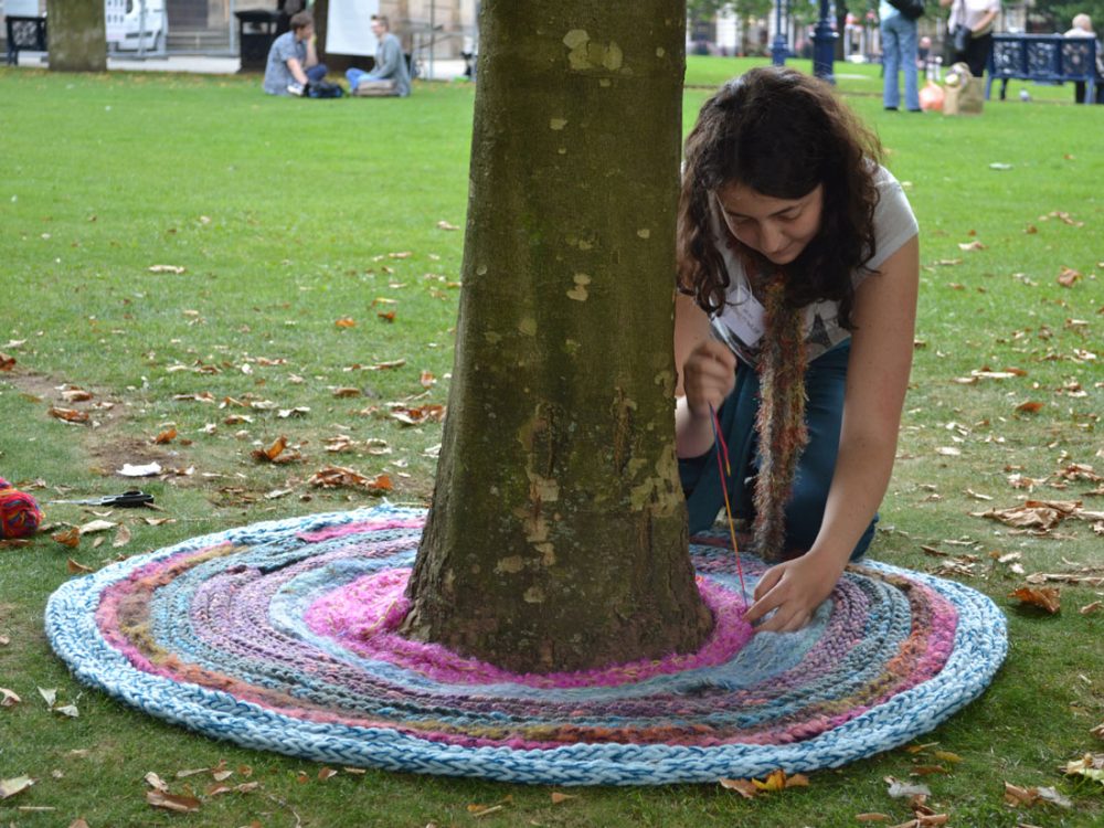 Sarah kneeling by a tree sewing together long strips of knitted wool to create a cirular shape around the bottom of the tree.