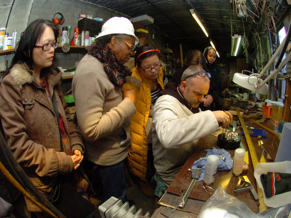 A group of women watch a jewellery maker at his workbench.