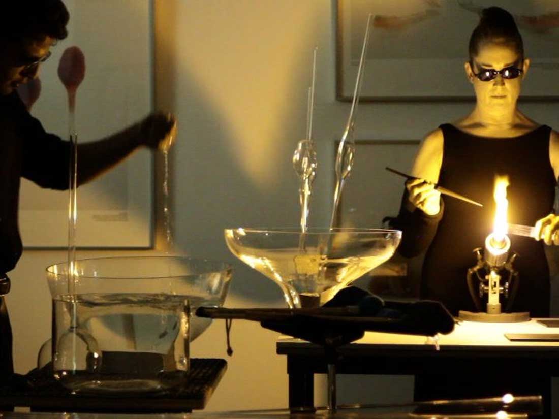 Inside a gallery a woman is performing with tools and glass, there is a large flame, other galss vesssels are filled with water.