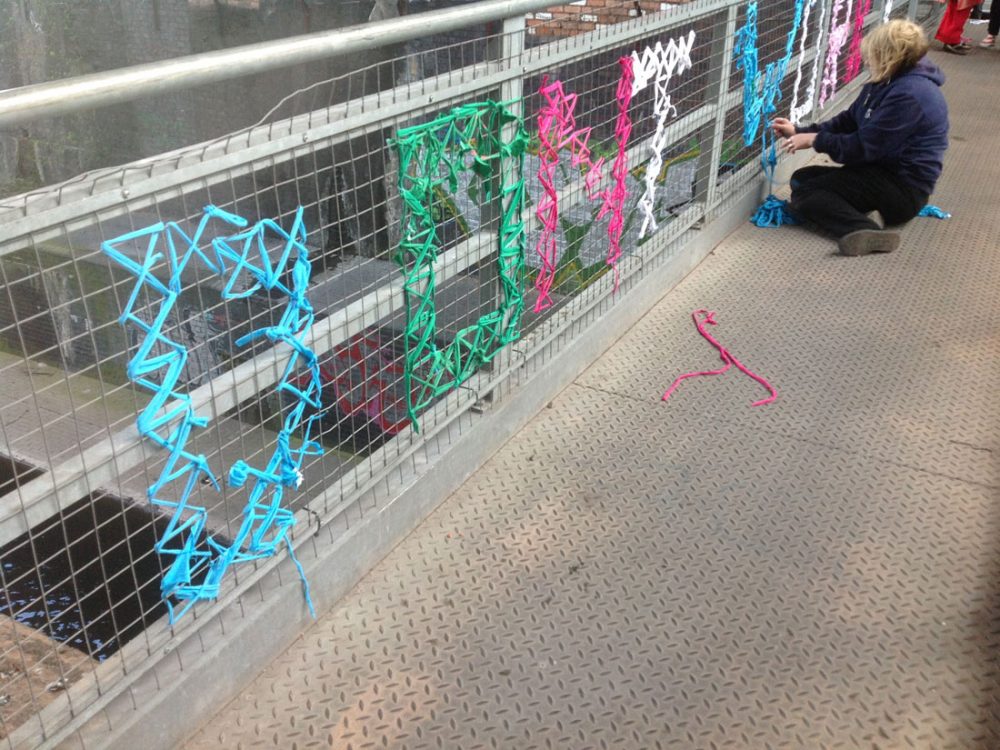 The words 'don't worry' are weaved onto a metal bridge using yarn of various colours. A lady sits and finishes weaving the letter W.