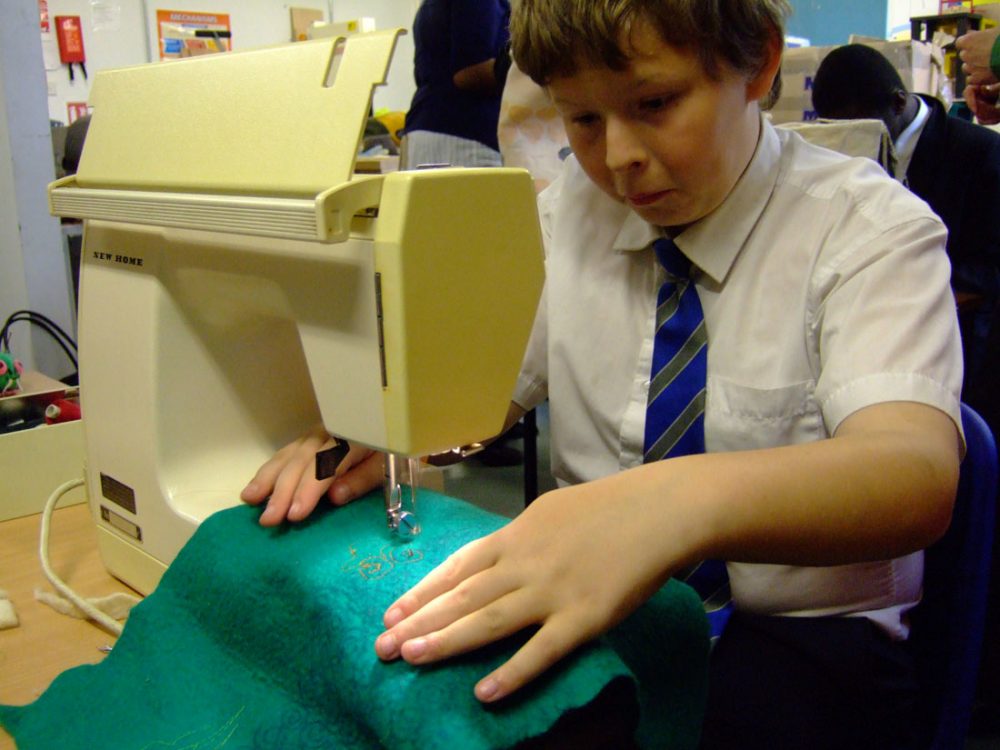 A young boy tentatively uses a sewing machine to sew an intricate design into a teal blue fabric.