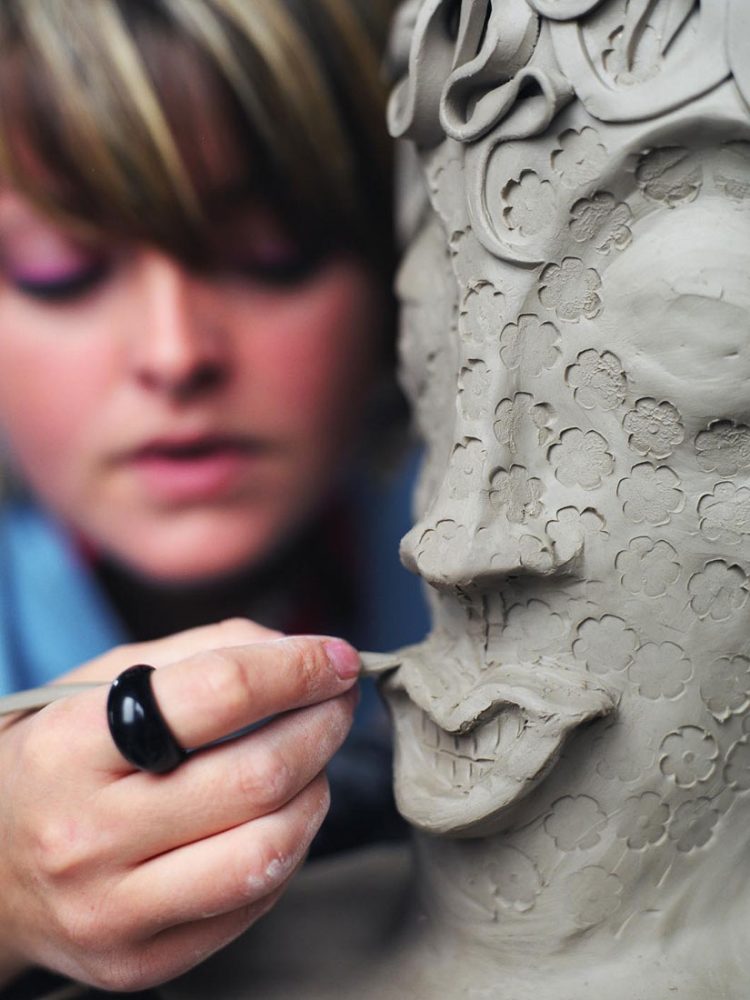 A close up of an artist carving lips into an intricate clay head.