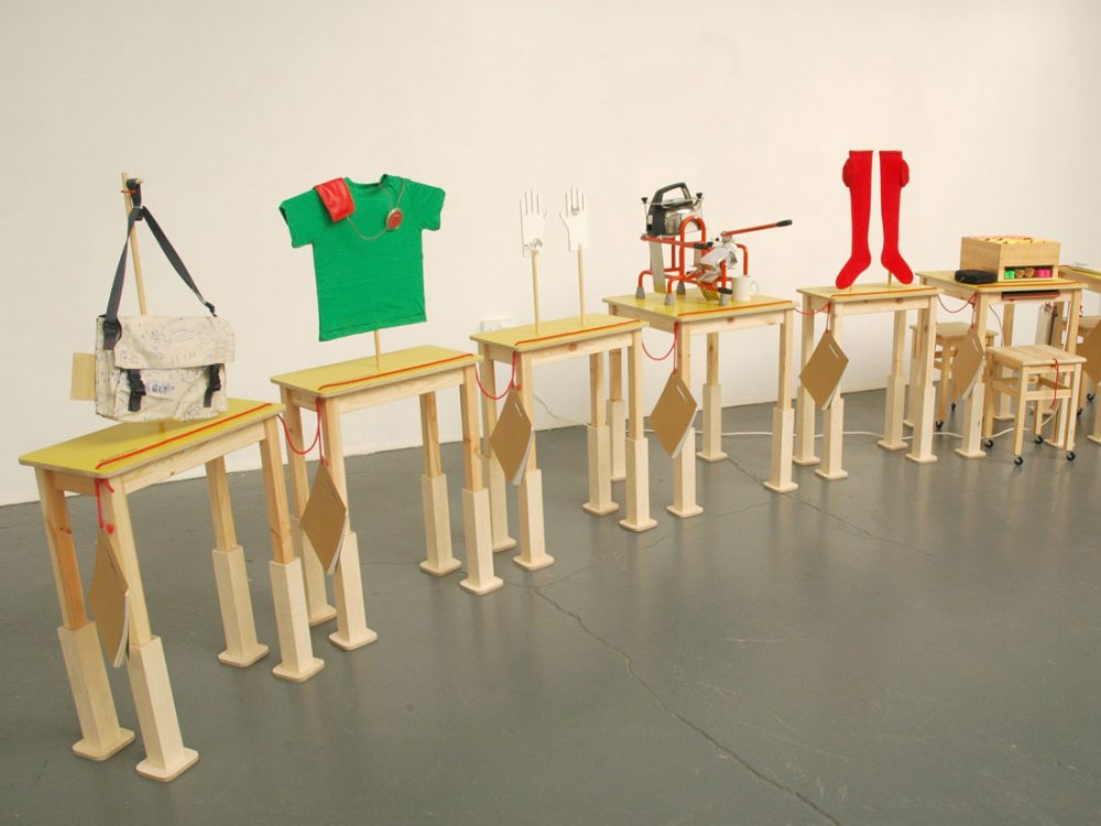 An exhibition with six small wooden tables with various objects placed on top of them including a bag.