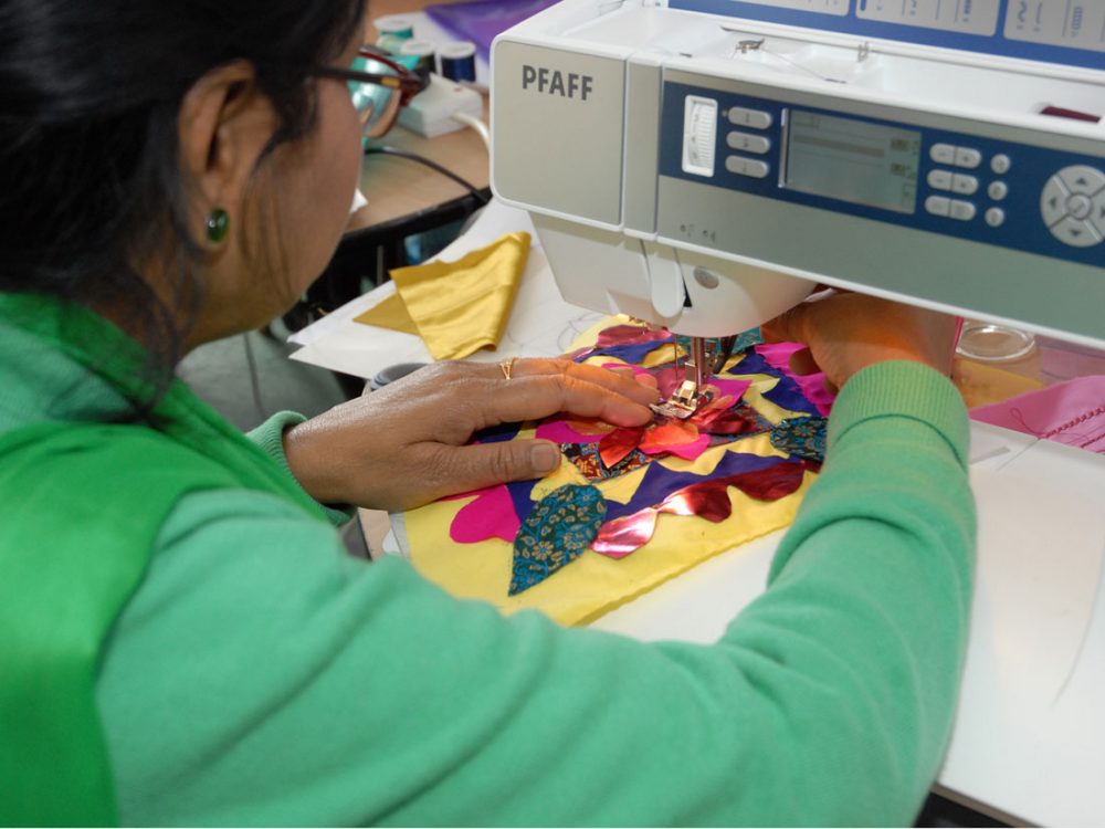 A woman stitches her colourful flower design on to yellow fabric using a sewing machine.