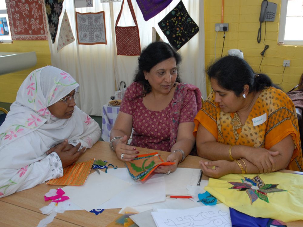 Three women sit at a table in deep discussion whilst holding various fabrics.
