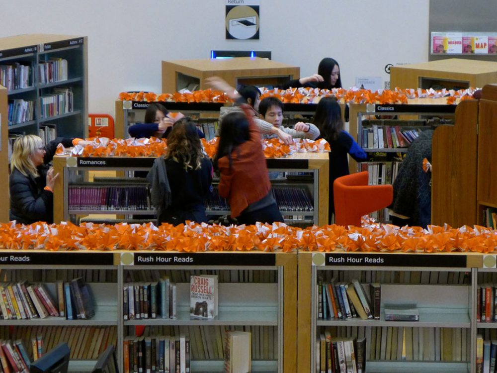 People in a library reach above the top shelf to pick up the origami shapes made from orange and white paper.