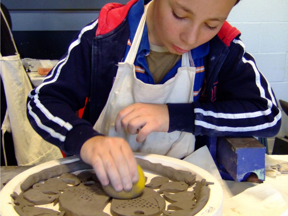 A young boy cleans up the side of the clay using a sponge.