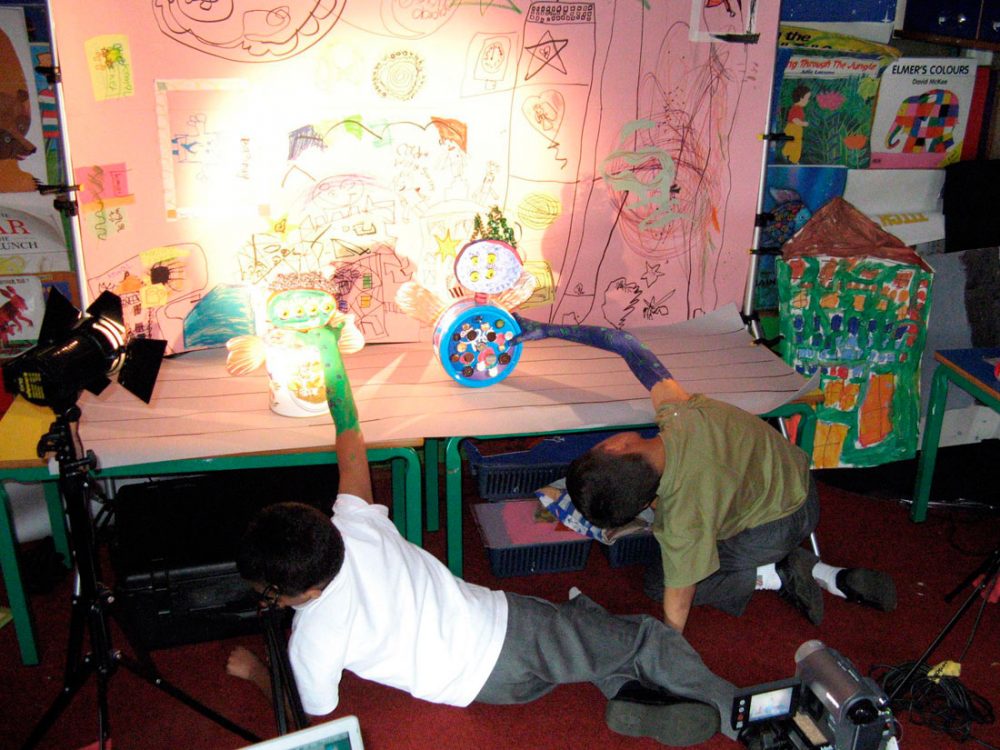 Two children use their figures made from found objects. They have drawn a backdrop setting and are using lighting.