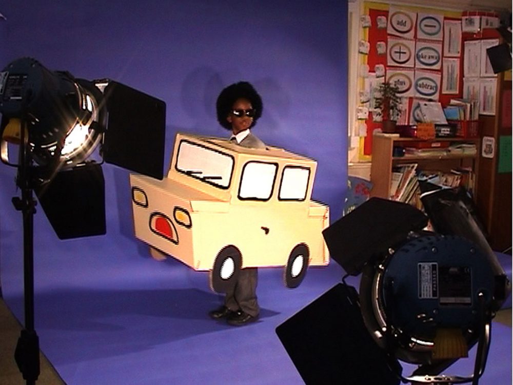 A young boy in sunglasses and a cardboard car stands infront of a blue screen with various lights on him.