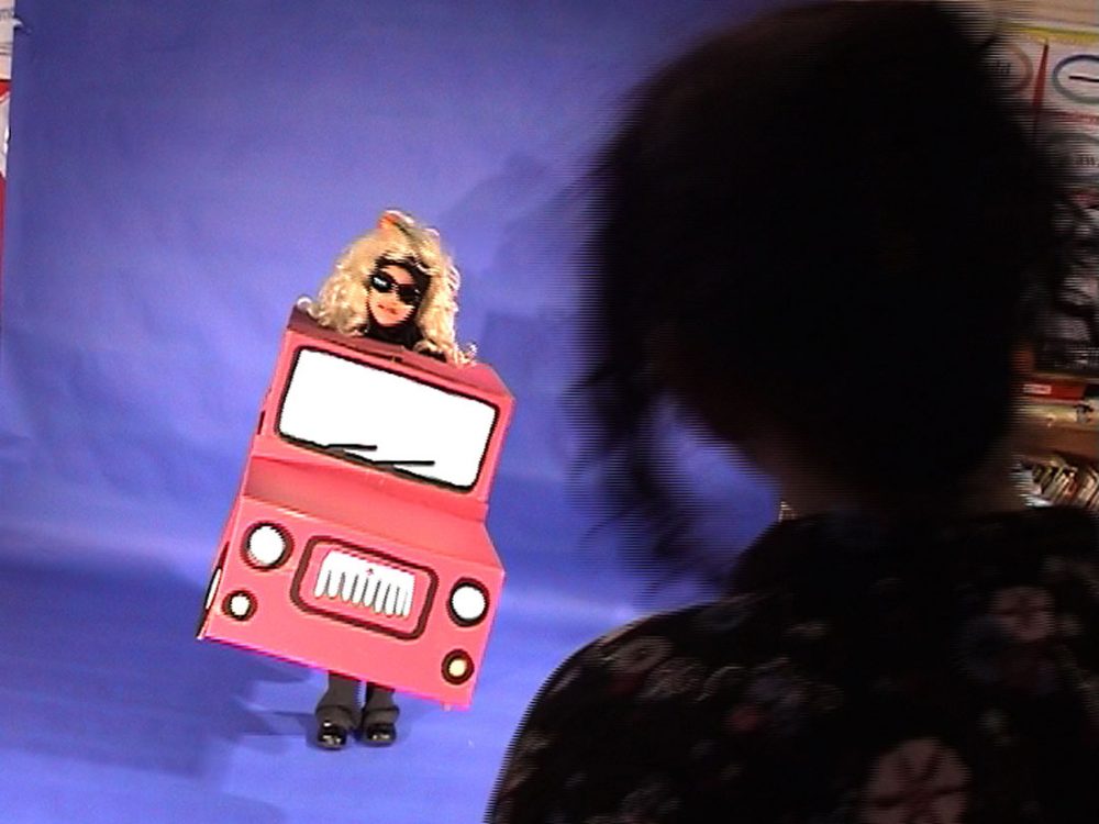 A child dresses in a cardboard box car with sunglasses and a blonde wig infront of a blue screen whilst a lady watches.