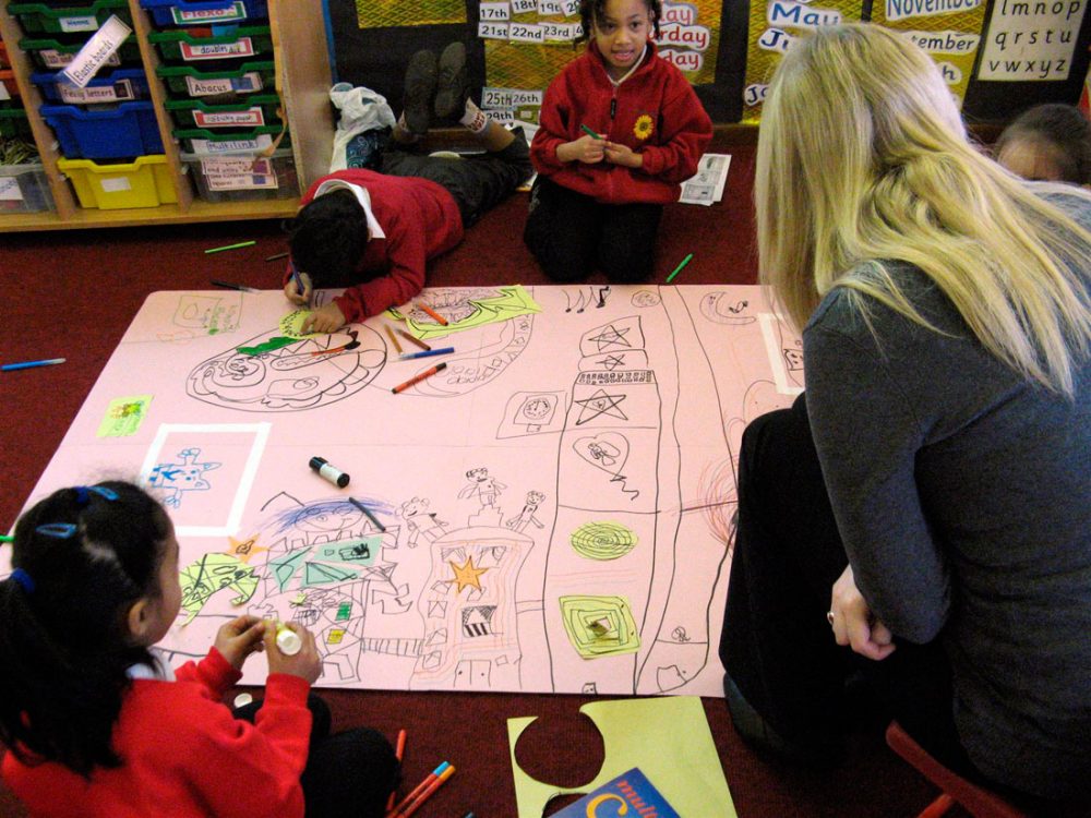 Three children draw on to a large piece of paper using pens whilst a lady supervises.