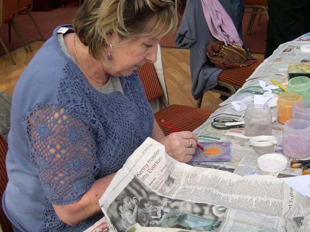 A woman sketches a design onto sheet metal with enamel powders placed on to the sheet metal.
