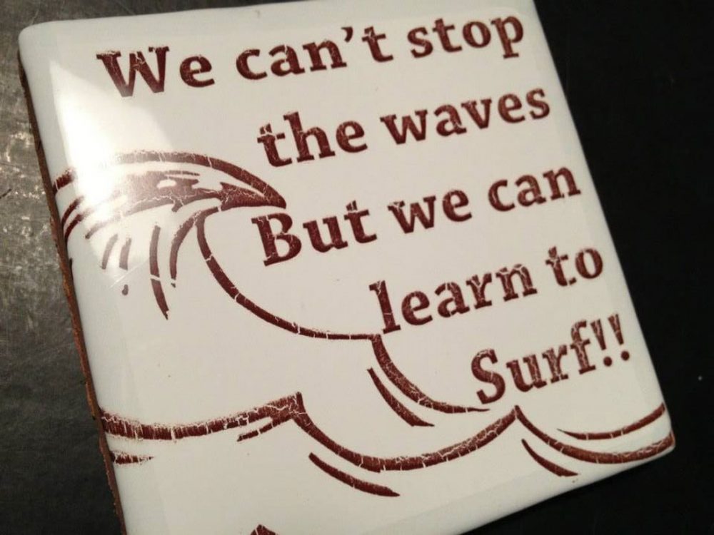 A white tile with brown text saying 'We can't stop the waves but we can to learn to surf!!' with a drawing of waves next to it.