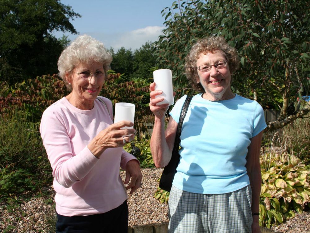 Two older women hold simple white glass cups whilst smiling.