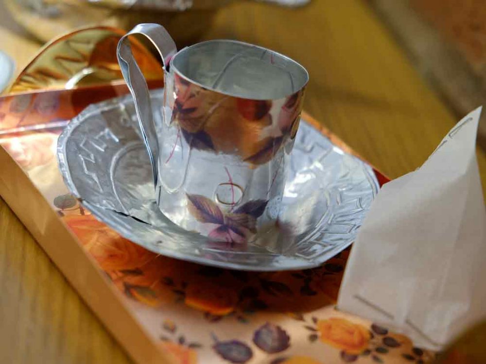A tea cup with a handle adn a small tray shaped from thin metal that has been printed with flowers and marked with indentations.