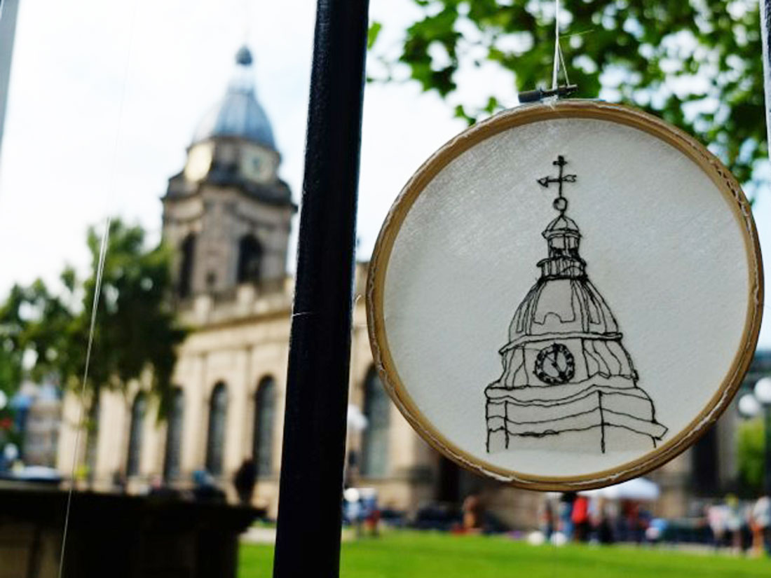 An emboidery ring hangs on the railings outside Birmingham Cathedral, the embroidery shows the clocktower of the same cathedral.