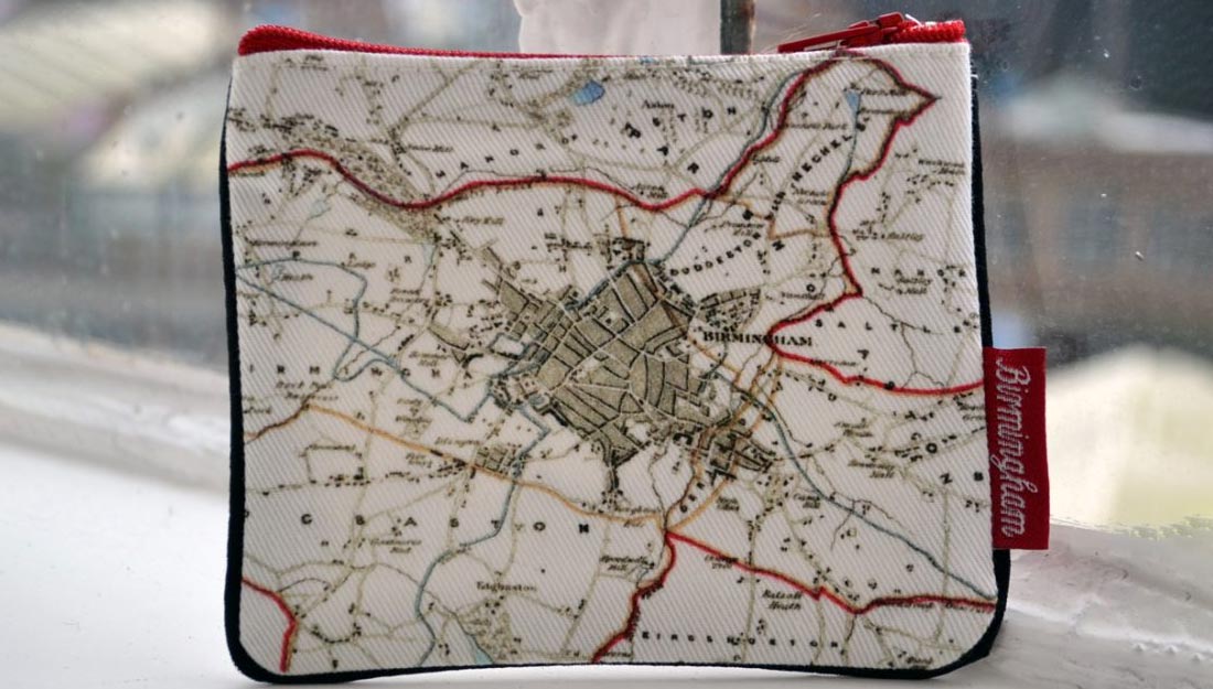 a purse sits on a windowsill. It is simple in design with an old map of Birmingham printed onto it and red trimmings. The tag says 'Birmingham'.