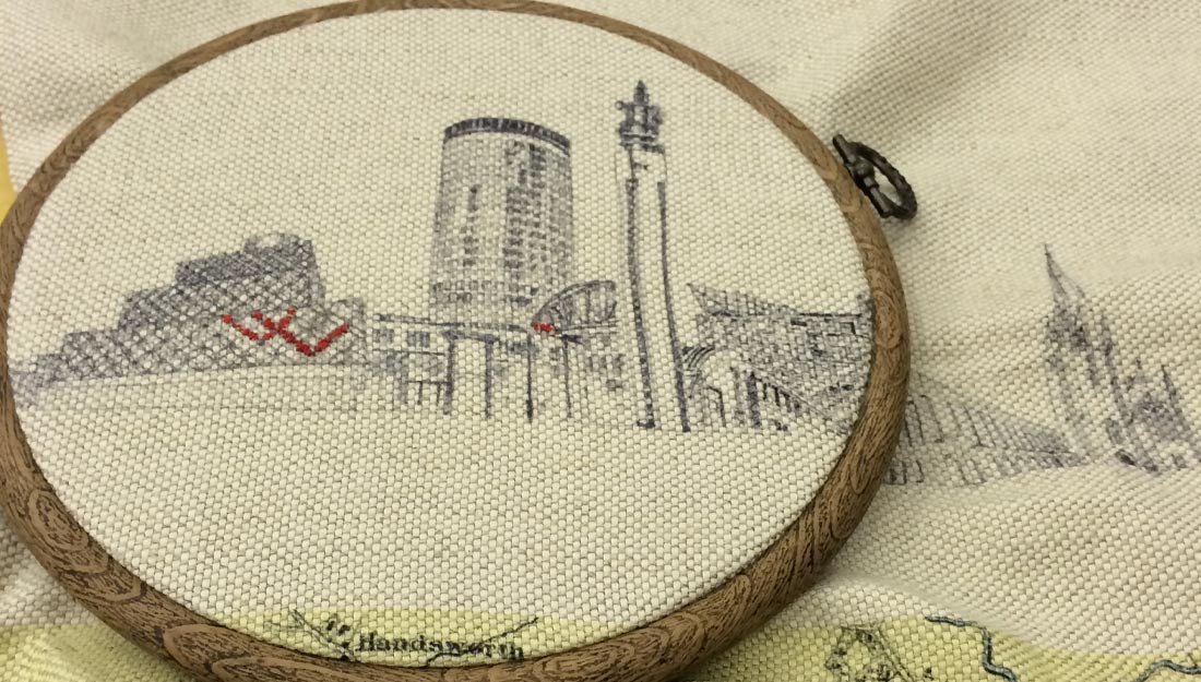 a cream coloured fabric with a printed illustration of a city skyline is stretched into a wooden embroidery hoop with some red embroidery stitches visible in the centre.