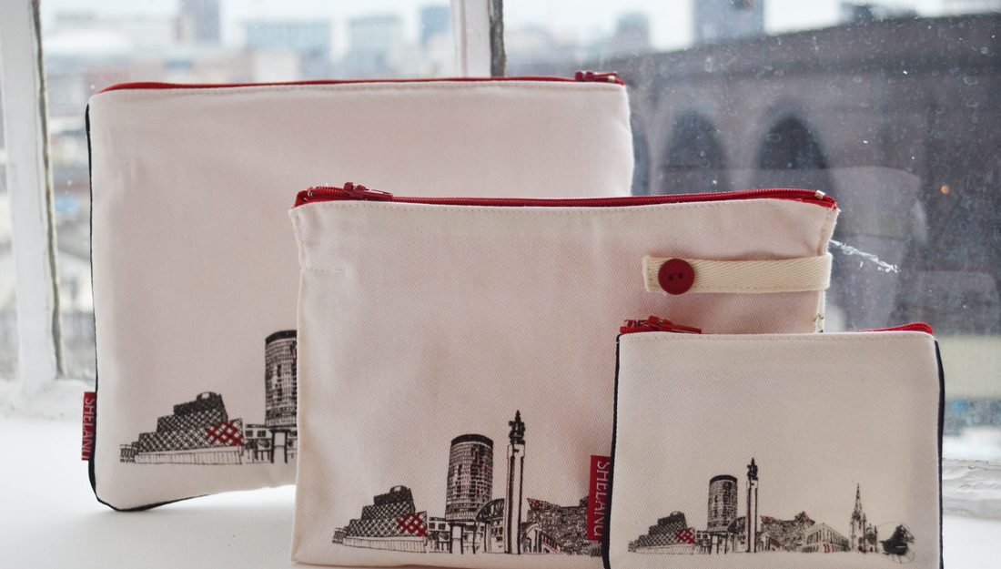 The i pad case, wash bag and purse sit on a windowsill. They are simple in design with a black and white line drawing of the skyline and red trimmings.