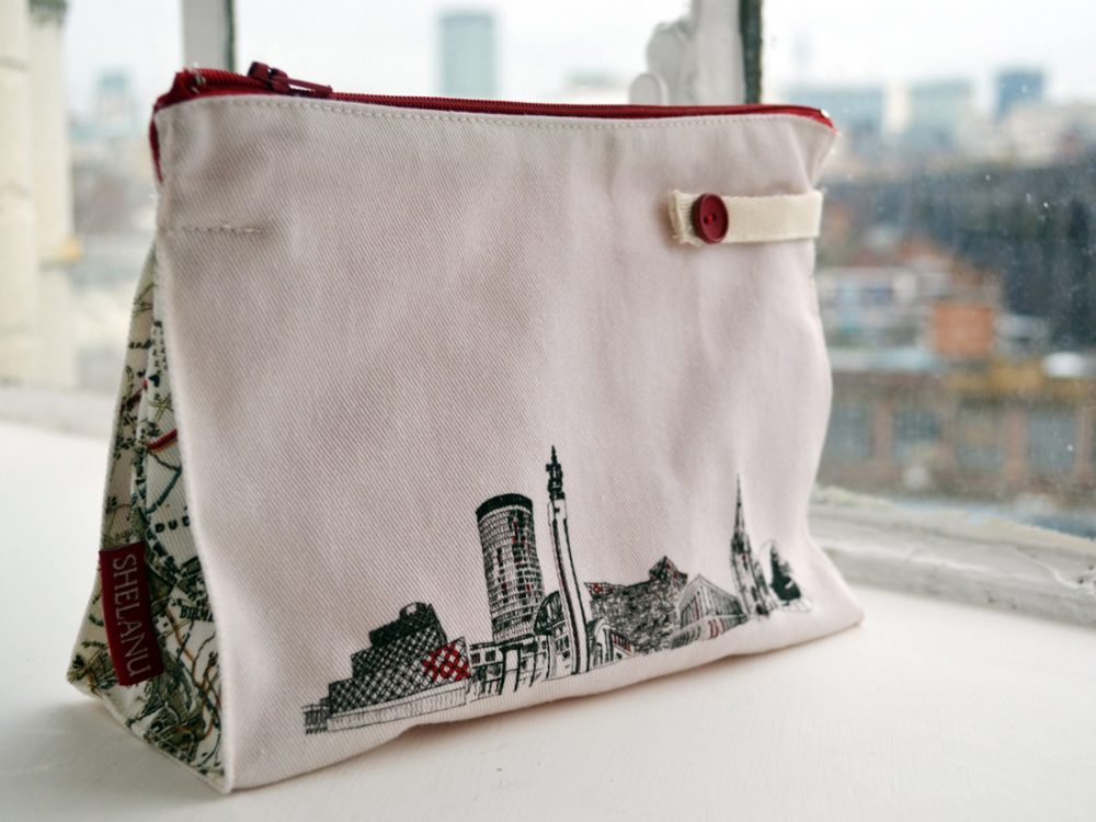 The wash bag sits on a windowsill. It is simple in design with a black and white line drawing of the skyline and red trimmings.