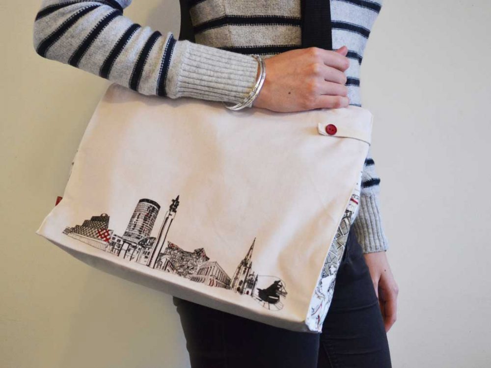 A young woman wears the bag over her shoulder. It is simple in design with a black and white line drawing of the skyline and red trimmings.