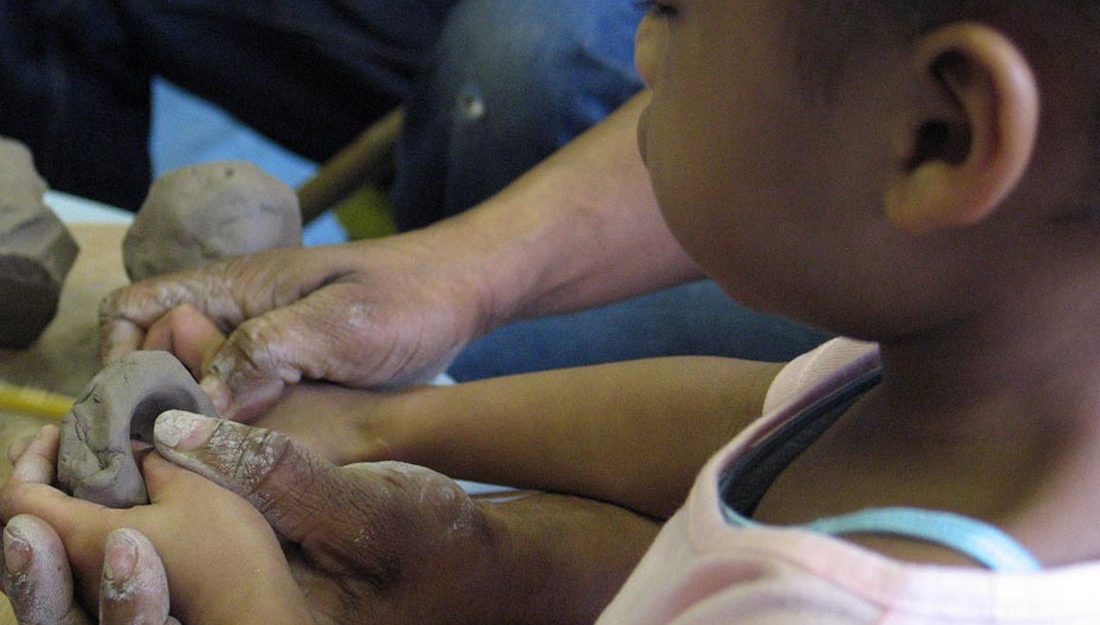 A small boy holds a pices of wet clay and pushes his fingers into it to make a small pot. The image shows adult hands working with the childs hands.