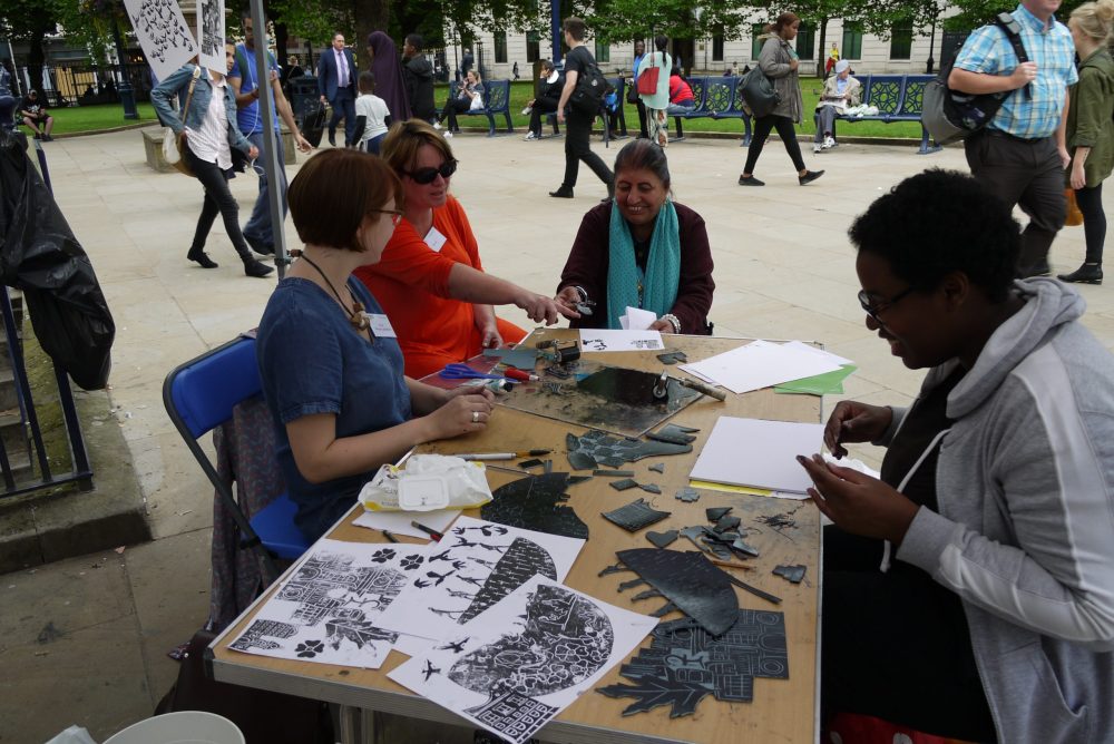 Four women of different ages are sat at a table in a public square making stencils. One woman is talking and pointing at something on the table and the other ones are laughing