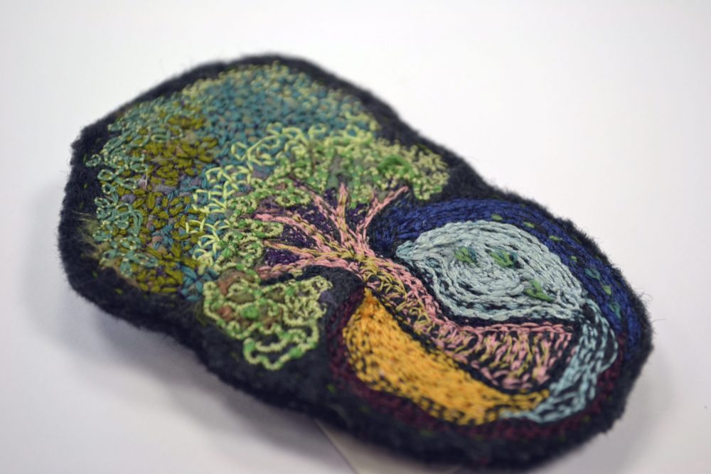 intricate embroidery patterns on felt of tree with surrounding patterns. colours include pale pink/green tree and background felt colour dark grey