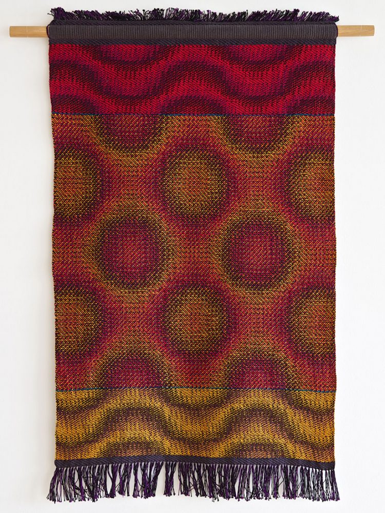 A woven piece of fabric of various colours hangs from a wall.
