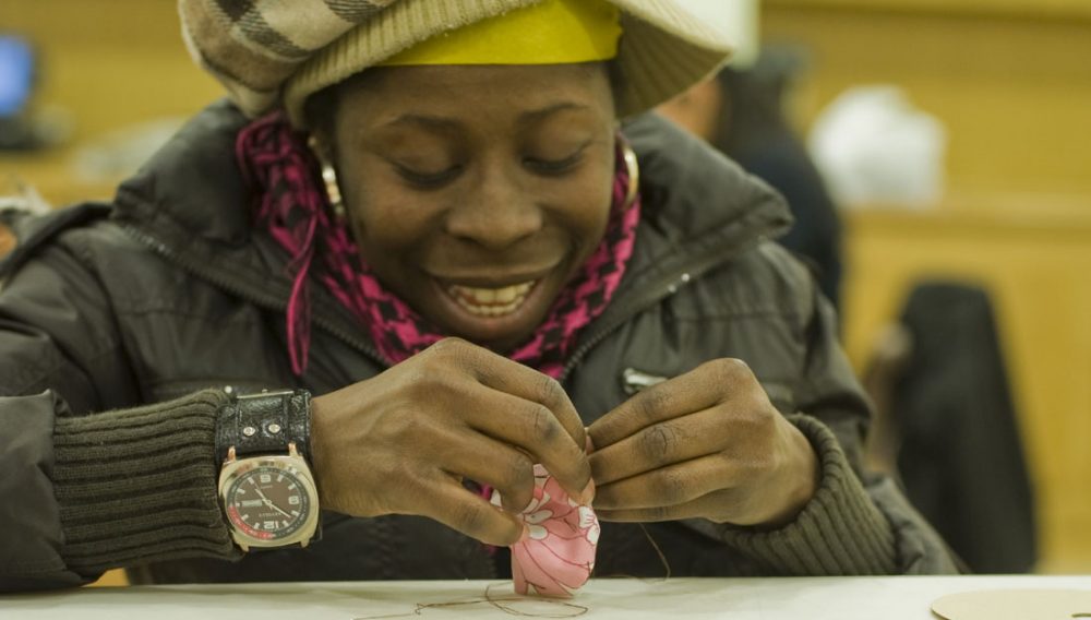 A lady smiles as she stitches into fabric using needle and thread.