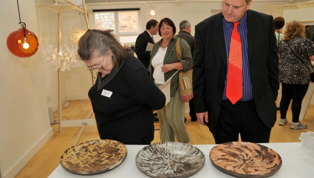 A man and woman admire three ceramic dishes on a table at an exhibition.