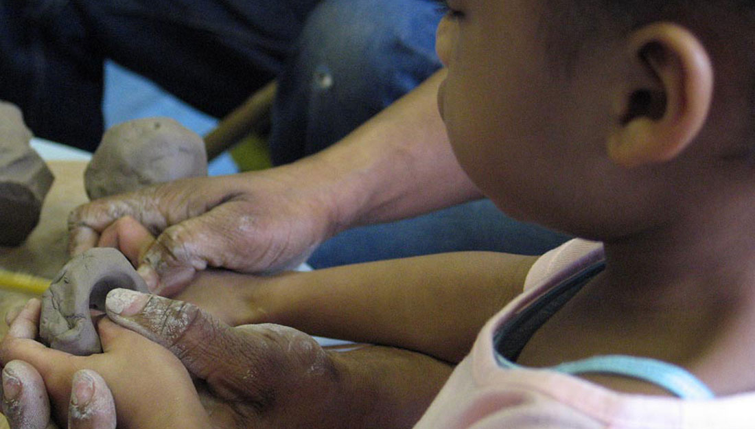 A small child and an adult hold a piece of wet clay and mould it together.